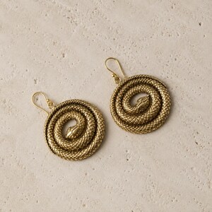 May include: A pair of gold-colored earrings featuring a coiled snake design. The earrings have a circular shape with a detailed texture, and a hook for easy wear. The snake's head is visible at the center of the coil.