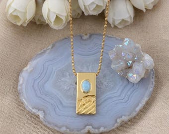 May include: A gold-colored pendant necklace with a rectangular pendant featuring a light blue oval stone. The pendant hangs from a delicate gold chain. The necklace is displayed on a blue and gray stone surface.