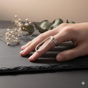 May include: A silver ring with a unique design. The ring features a long, oval-shaped element that extends over the finger, connected to two bands that encircle the base of the finger. The ring is displayed on a hand against a neutral background.