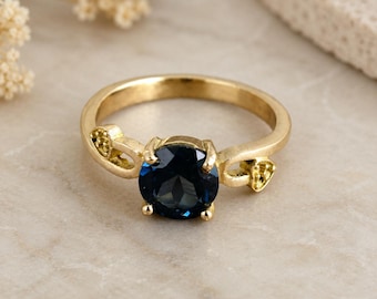 May include: A gold ring with a large, round, dark blue gemstone. The ring has decorative accents on the band and around the gemstone setting. The ring is set against a neutral background.