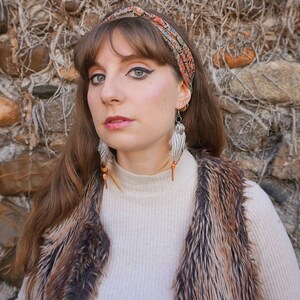 May include: A woman wearing a patterned headband, feather earrings, and a faux fur vest. The earrings have white feathers, wooden beads, and metal accents. The vest is brown and black. She is wearing a cream-coloured turtleneck.
