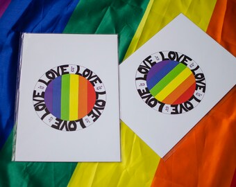 May include: Two white cards with a rainbow circle design and the text "Love is Love" printed around the circle. The cards are on top of a rainbow colored fabric.