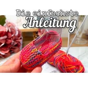 May include: A close-up of a colourful knitted baby shoe with a clear plastic form inside. The shoe is made with a multi-coloured yarn in shades of pink, orange, yellow, and purple. The yarn is also visible in a ball next to the shoe.