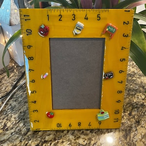 May include: A yellow wooden picture frame with ruler markings and decorative school-themed magnets. The frame has a brown interior and is decorated with magnets of an apple, pencil, and a small chalkboard.