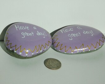 May include: Two painted purple rocks with gold accents and white polka dots. Each rock has the words "Have a great day!" written in white.