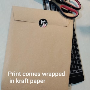 May include: A brown kraft paper envelope sealed with a "Thank You" sticker featuring floral accents. The text "Print comes wrapped in kraft paper" is visible. A pair of scissors and a paper cutter are in the background.