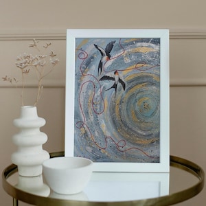May include: A framed abstract painting featuring two swallows in flight against a swirling background of gray, blue, and gold. Red lines weave through the composition. The white frame sits on a mirrored table.