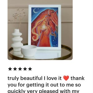 May include: A framed art print featuring a woman with long red hair and a horse, set against a blue starry sky. The woman holds a glowing object. The artwork is displayed on a table with a white vase and bowl.