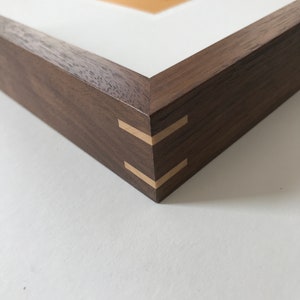 Puede incluir: Primer plano de un marco de fotos de madera con acabado marrón oscuro. El marco tiene un paspartú blanco y un interior naranja claro. Las esquinas están unidas con una incrustación decorativa de color claro. El marco está sobre un fondo blanco.