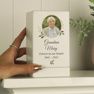 May include: White ceramic memorial box with a photo of a woman and the text "Grandma Mary Forever in our Hearts 1945 - 2025".