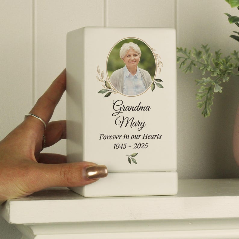May include: White ceramic memorial box with a photo of a woman and the text "Grandma Mary Forever in our Hearts 1945 - 2025".
