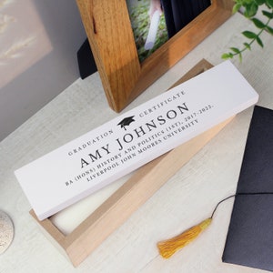 May include: A white wooden box with a white plaque on top. The plaque has a black graduation cap icon and the text "GRADUATION CERTIFICATE AMY JOHNSON BA (HONS) HISTORY AND POLITICS (1ST), 2017-2022. LIVERPOOL JOHN MOORES UNIVERSITY".