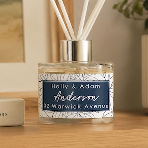 May include: A clear glass reed diffuser bottle with a silver cap and white reeds. The label features a dark blue rectangular centre with the text Holly & Adam, Anderson, and 32 Warwick Avenue, surrounded by a white border with a black leaf pattern.