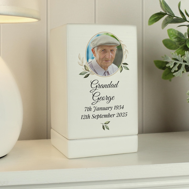 May include: White wooden box with a photo of a man in a cap. The box has a gold trim and a floral design. The text on the box reads "Grandad George 7th January 1934 12th September 2025".
