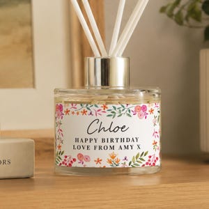 May include: A clear glass reed diffuser with a silver collar and white reeds. The label features a colourful floral border with the text "Chloe" and "HAPPY BIRTHDAY LOVE FROM AMY X" printed in black ink on a white background.