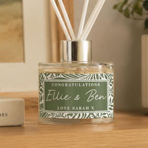 May include: A clear glass reed diffuser with a silver collar and white reeds. The label is sage green with a white botanical border and the text: CONGRATULATIONS, Ellie & Ben, LOVE SARAH X. The diffuser contains a light-coloured liquid fragrance.