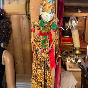 Puede incluir: Una marioneta de varilla Wayang Golek con rostro blanco y rasgos pintados, un tocado alto puntiagudo en rojo y dorado, y una falda larga con patrón geométrico marrón y beige. La marioneta tiene brazos articulados y detalles de tela verde.