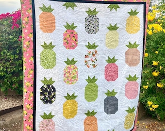May include: A handmade quilt featuring a repeating pattern of colorful pineapple shapes on a white background. The pineapples are various colors and patterns, with green leafy tops. The quilt has a pink floral border.
