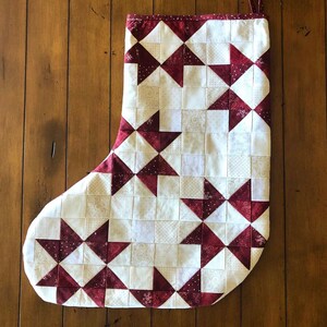 Peut inclure: Une chaussette de Noël faite à la main avec un motif patchwork. La chaussette présente un motif d'étoiles bordeaux et blanches, avec un fond crème. Le tissu a une texture matelassée et la chaussette est présentée sur une surface en bois.