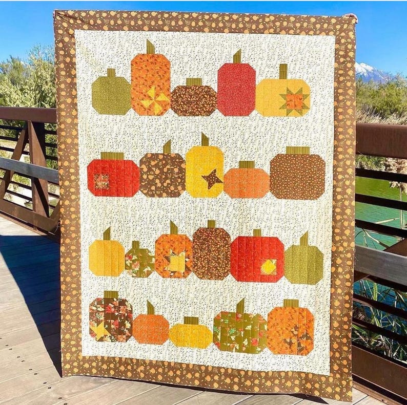 May include: A quilted wall hanging featuring a repeating pattern of stylized pumpkins in shades of orange, yellow, brown, and green. The pumpkins are arranged in rows on a cream background, with a brown border.