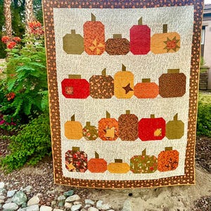 May include: A handmade quilt featuring a repeating pattern of colorful pumpkins in shades of orange, red, brown, and green. The pumpkins have various patterns and textures, set against a cream background with a brown border. The quilt is displayed outdoors.
