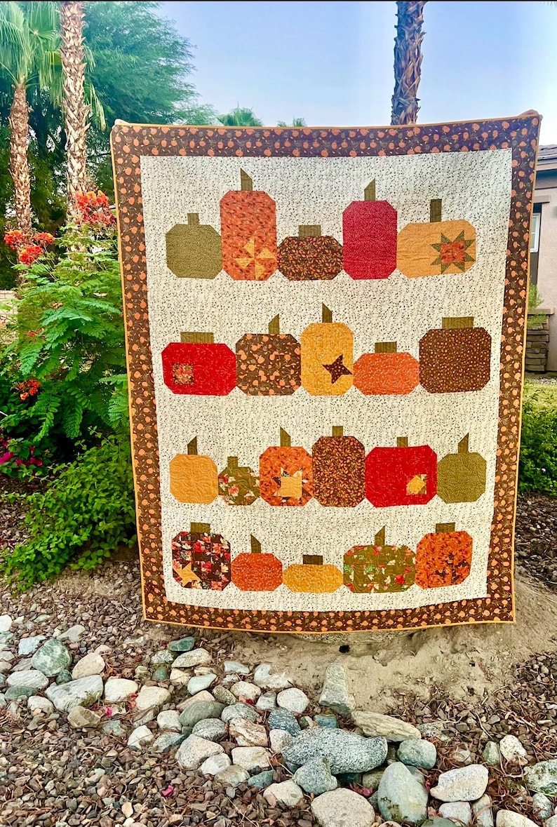 May include: A handmade quilt featuring a repeating pattern of colorful pumpkins in shades of orange, red, brown, and green. The pumpkins have various patterns and textures, set against a cream background with a brown border. The quilt is displayed outdoors.