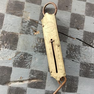 May include: Vintage brass hanging scale with a ring at the top and a hook at the bottom. The scale has a vertical measurement indicator with numbers and markings. The scale is marked "PATENTED" and "SPRING BALANCE".