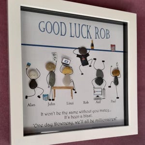 May include: A white framed artwork with the text "GOOD LUCK ROB" in blue. The artwork depicts pebble stick figures, each representing a person. The text "It won't be the same without you matey..." is also included.