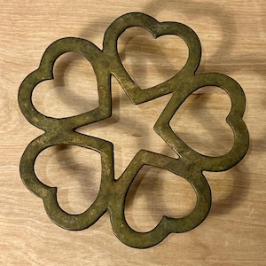 May include: A bronze-colored trivet with a star-shaped center and six heart-shaped cutouts. The trivet is made of metal and has a rustic, aged appearance. It is designed to protect surfaces from hot cookware.