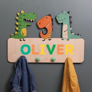 May include: Wooden coat rack with three hooks featuring a green, orange, and green dinosaur design. The rack has the name "OLIVER" written in colorful letters.