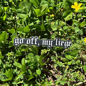 May include: A black and white sticker with the text "go off, my liege" in a gothic font. The sticker is placed on a bed of green clover.