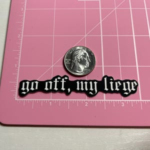 May include: Black and white sticker with the text "go off, my liege" in a gothic font. The sticker is on a pink cutting mat with a US quarter for scale.