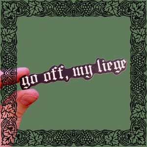 May include: A black sticker with white lettering that reads "go off, my liege" in a gothic font. The sticker is held in a hand against a green background.