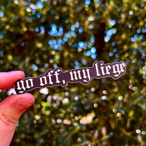 May include: A black sticker with white text that reads "go off, my liege" in a gothic font. The sticker is held in a hand against a blurry background of green and yellow foliage with out-of-focus lights.