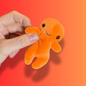 May include: A small, bright orange plush toy shaped like a simple humanoid figure. The soft, fuzzy character features two round, black plastic eyes and a thin, stitched black line for a mouth, with short arms and legs extending from the body.