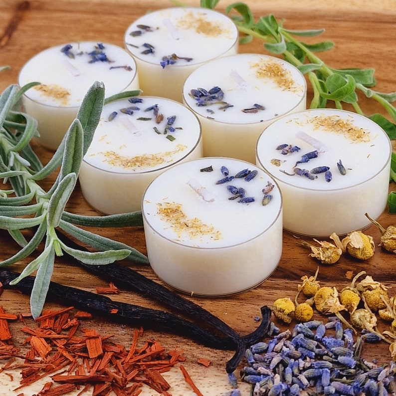 May include: A set of six white tea light candles with dried lavender and chamomile flowers on a wooden surface. The candles are surrounded by dried herbs and flowers.
