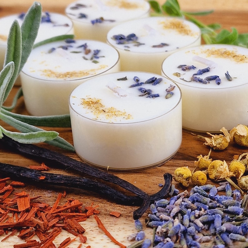 May include: A set of six white tea light candles with dried lavender and chamomile flowers on a wooden surface. The candles are in glass holders and are surrounded by dried herbs and flowers.