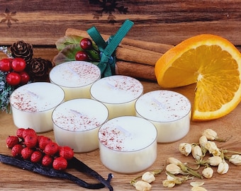 May include: Six cream-coloured tea light candles sprinkled with spice, alongside cinnamon sticks, an orange slice, red berries, and vanilla beans. The arrangement is set on a wooden surface, creating a warm and inviting atmosphere.