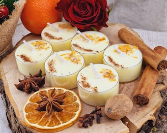 May include: Cream-coloured tea light candles with orange zest and cinnamon, displayed on a wooden surface with a red rose, orange, star anise, and cinnamon sticks. The candles have white wicks.