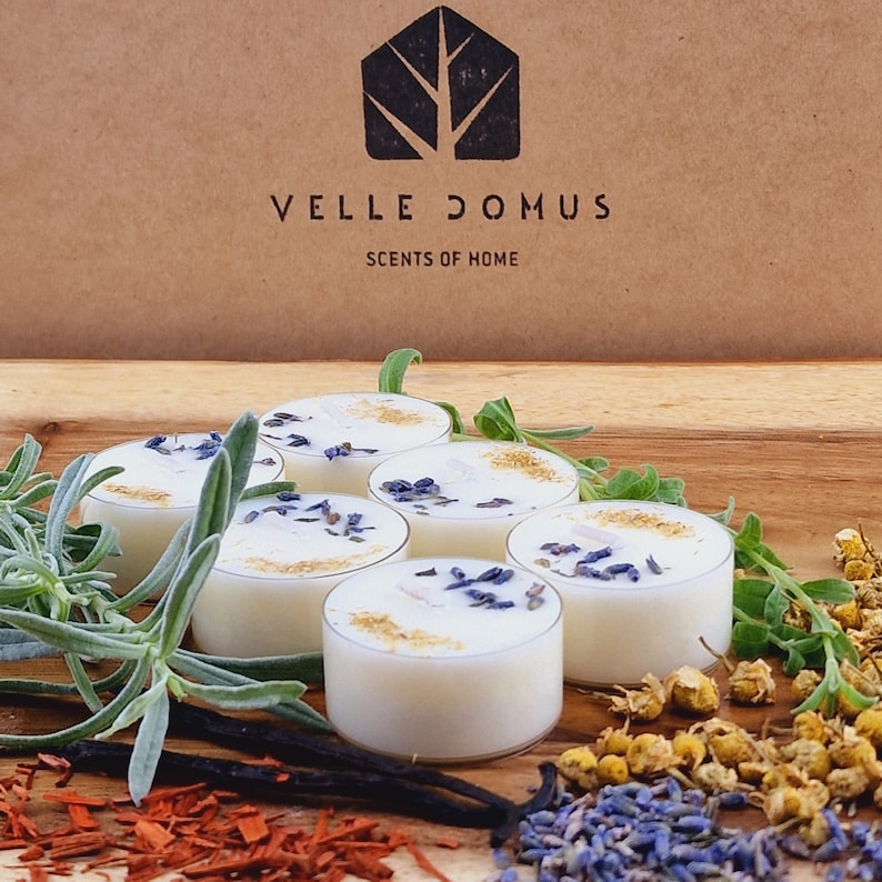 May include: Six white tea light candles with dried lavender and gold flakes on top. The candles are arranged in a circle on a wooden surface with dried herbs and spices around them. The text "VELLE DOMUS SCENTS OF HOME" is visible on a brown box in the background.