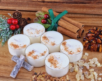May include: A selection of tea light candles with a spice topping, cinnamon sticks, and amber beads on a wooden surface. Decorative elements include pine cones, berries, and a green ribbon.