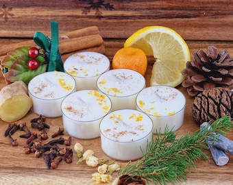 May include: Six white tea light candles with orange zest and cinnamon, alongside cinnamon sticks, cloves, ginger, a lemon slice, a mandarin orange, and pine cones on a wooden surface. A festive display.