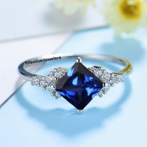 May include: A silver ring with a square blue sapphire gemstone surrounded by smaller clear gemstones. The ring is set on a blue background.