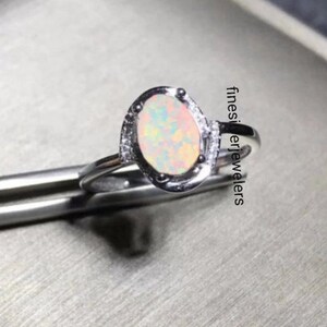 Pode incluir: Um anel de prata com uma gema de opala branca engastada em uma moldura de prata. O anel é cercado por uma fileira de pequenas gemas transparentes.