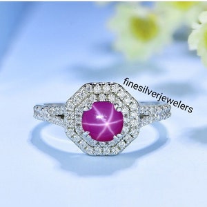 May include: A silver ring with a pink star ruby gemstone surrounded by a double halo of smaller diamonds. The ring is set in a octagonal shape.