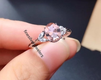 May include: A silver ring with a pink oval gemstone and smaller clear gemstones set in a leaf design. The ring is on a finger.