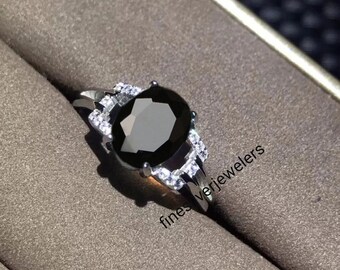May include: A silver ring with a large oval black gemstone surrounded by smaller white gemstones. The ring is set in a white metal band. The text "fines verjewellers" is visible on the ring.