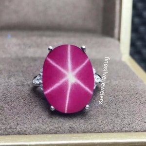 May include: A silver ring with a large oval-shaped pink star ruby gemstone surrounded by smaller diamonds. The ring is set in a white metal band.