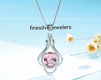 May include: A silver necklace with a pink oval gemstone pendant. The pendant is set in a delicate silver frame with smaller clear gemstones. The necklace is on a white background.