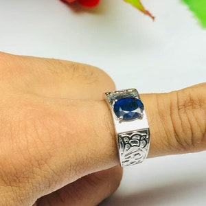 May include: A silver ring with an oval-shaped blue gemstone set in the center. The ring band is decorated with an intricate design.